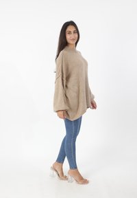 Beige oversized trui met ribbelstructuur, hoge hals en verlaagde schouders, gecombineerd met blauwe skinny jeans en heldere blokhak sandalen.