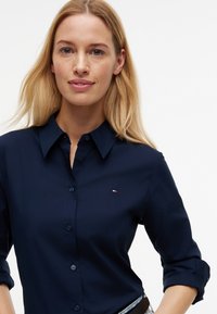 Femme blonde portant une chemise bleu marine boutonnée avec les manches retroussées au-dessus des coudes, debout devant un fond blanc uni.