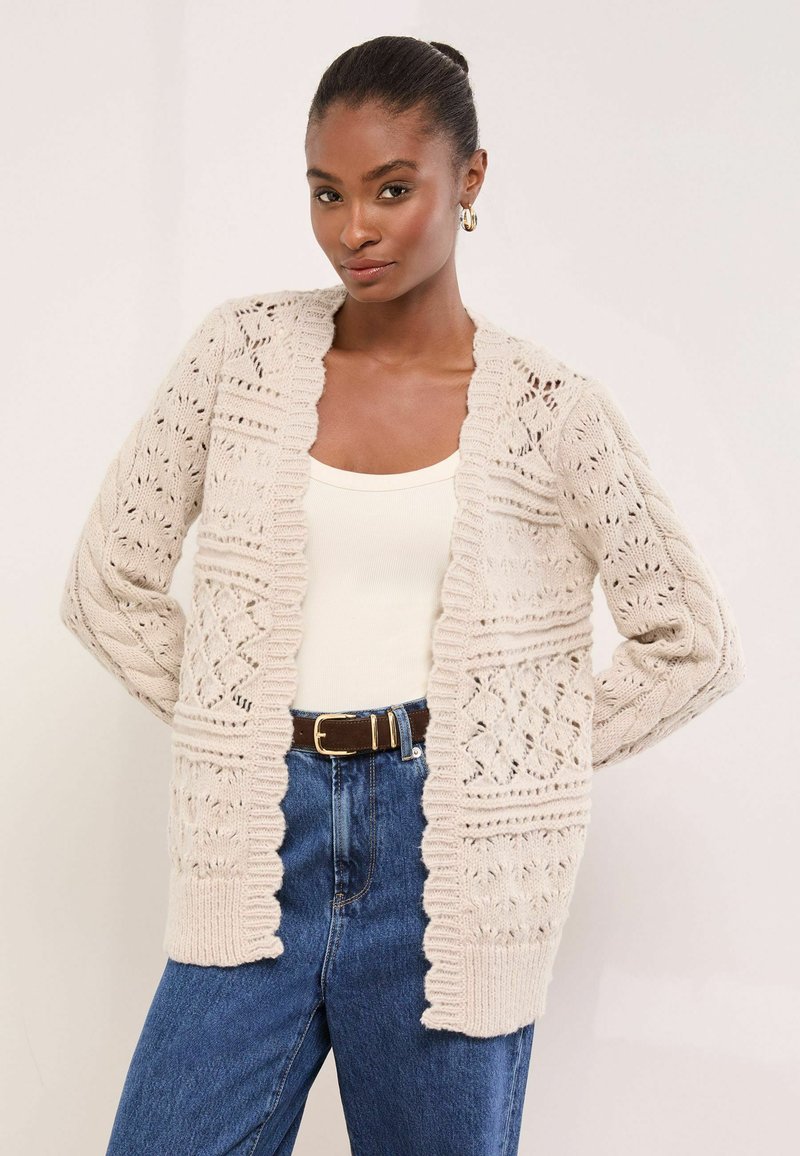 Beige kötött kardigán bonyolult nyitott mintákkal, bordázott szélekkel és hosszú ujjakkal, világos trikó és magas derekú farmernadrág felett viselve.