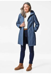 Blauwe waterdichte jack met een capuchon, voorrits, zijzakken en verstelbare trekkoorden, gecombineerd met donkere spijkerbroeken en bruine enkelboots.