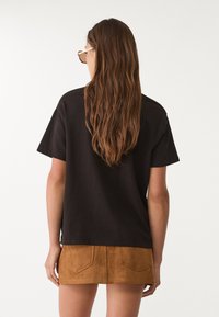 Czarna bawełniana koszulka z krótkim rękawem w połączeniu z mini spódniczką z tanowego sztucznego zamszu, posiadającą dwie tylne kieszenie. Widoczne długie, falujące brązowe włosy.