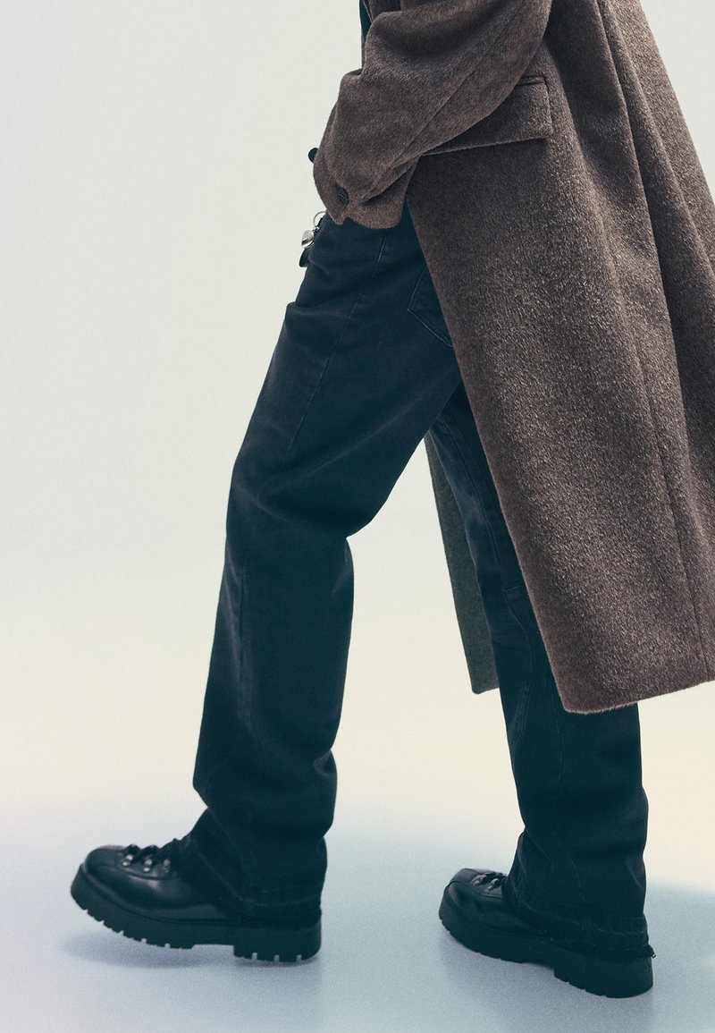 Black lace-up shoes with chunky soles, paired with dark loose-fitting pants and a textured brown overcoat, creating a layered look.