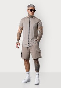 Chemise beige à manches courtes avec un détail de rayure verticale noire. Associée à un short cargo beige avec des poches latérales. Baskets blanches et lunettes de soleil.