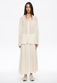Camisa abotonada de manga larga, ligera y transparente, en color blanco roto, combinada con una falda maxi fluida blanca rota y zapatillas plateadas.