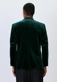 Green velvet blazer with a fitted design, single vent at the back, and three-button cuffs, featuring a smooth texture and elegant silhouette.