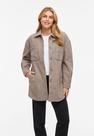 Smilende kvinde med langt blondt hår iført en taupefarvet skjortejakke med knapper over en hvid top og sorte bukser, med hånden i lommen.