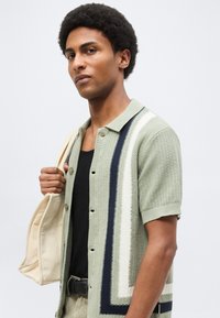 Νέος άνδρας με αφρο μαλλιά, φοράει πράσινο ριγέ κοντομάνικο πουκάμισο, μαύρο αμάνικο και κουβαλά μια μπεζ τσάντα ώμου.