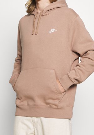 Beżowa bluza Nike z kapturem, z przednią kieszenią i sznurkami, noszona przez osobę z rękami w kieszeniach, w połączeniu z jasnymi spodniami.
