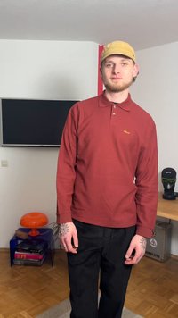 Polo rouge à manches longues côtelé avec col et patte de boutonnage, associé à un pantalon noir. Contient des tatouages visibles sur les mains.