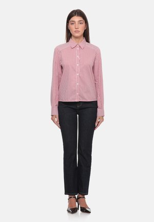 Donna in piedi di fronte che indossa una maglia a righe rosse e bianche a maniche lunghe, jeans scuri e scarpe nere con tacco su sfondo bianco.