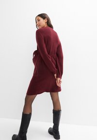 Bordeauxrotes gestricktes Wickelkleid mit langen Ärmeln, Gürtelschlaufe in der Taille und einer Länge bis zur Mitte des Oberschenkels, kombiniert mit schwarzen kniehohen Stiefeln.