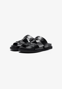 Zwarte leren sandalen met brede, gekruiste banden en gespdetails. De zool is plat en heeft een gestructureerde oppervlakte voor grip.