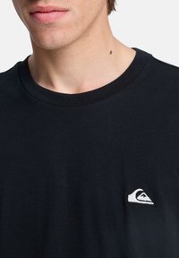 Camiseta de algodón negra con cuello redondo y mangas cortas, con un pequeño logotipo bordado en blanco en el pecho.
