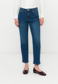 Person med blå denim jeans og burgunder patent lær flats, stående mot en ensfarget hvit bakgrunn.
