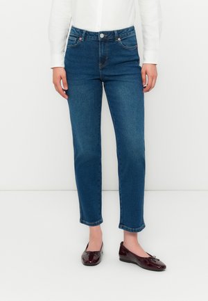 Persona vestida con jeans de mezclilla azul y planos de cuero lustrado burdeos, de pie contra un fondo blanco liso.