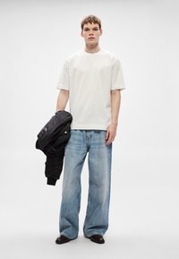 Tricou alb de bumbac, croială lejeră; blugi albaștri din denim supradimensionați; jachetă bomber neagră; model care ține jacheta, poartă pantofi închiși la culoare; fundal simplu.