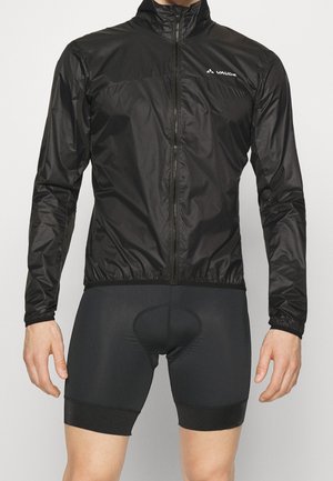 Man draagt een zwarte Vaude lichte jas met rits en zwarte gevoerde fietsbroek, staand tegen een effen achtergrond.
