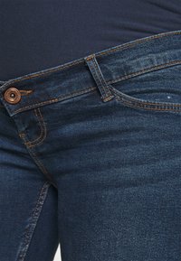 Tmavě modré denimové džíny s přiléhavým střihem, s oranžovým stehem a předním zapínáním na knoflík v pase. Hladká textura bez vzorů.