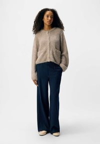 Hellbrauner, flauschiger Cardigan mit vorderen Taschen, Knopfverschluss und navyblauen, gestreiften, weit geschnittenen Hosen. Das Outfit hat eine lässige Passform und einen weichen Stoff.