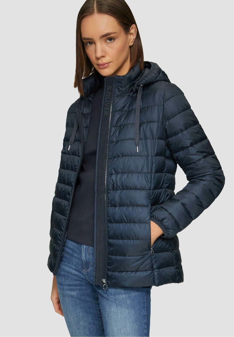 Veste réversible bleu marine avec un design matelassé horizontal, fermeture éclair, capuche ajustable et poches latérales. Matériau doux et léger.