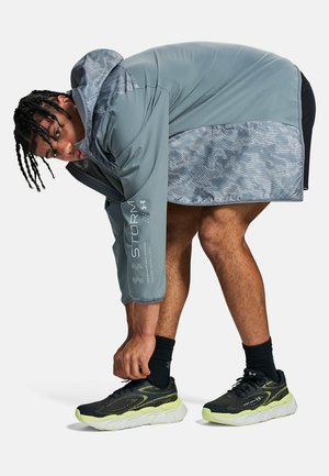 Mann beugt sich vor und bindet schwarze und neon-gelbe Laufschuhe, trägt eine graue wetterfeste Jacke und schwarze Shorts vor weißem Hintergrund.