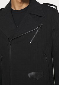 Närbild av en person som bär en svart motorcykeljacka med silverdragkedjor och ett texturerat mönster över en svart t-shirt med rund hals.