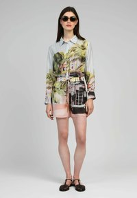 Femme portant des lunettes de soleil, une chemise à manches longues avec des boutons et un short à imprimé tropical et voiture vintage, des sandales tissées noires, se tenant contre un fond gris.