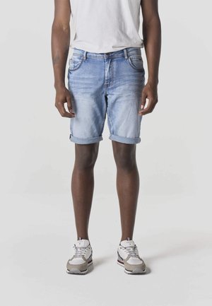 Persona vistiendo pantalones cortos de mezclilla azul claro remangados, zapatillas blancas y una camiseta blanca, de pie contra un fondo liso.