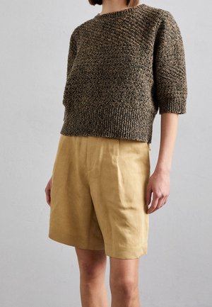 Kobieta w brązowym, teksturowanym, dzianym sweterku z rękawami do łokci i wysokim stanem beżowych szortów na jednolitym tle.