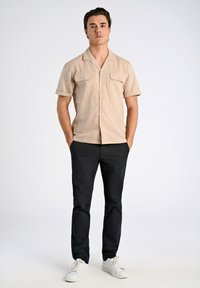 Lichtbeige T-shirt met korte mouwen en twee voorzakken, gecombineerd met donkerblauwe broek en witte sneakers, voor een effen achtergrond.