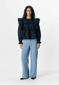 Blouse noire à volants avec un col rond et des volants superposés. Associée à un jean bleu clair à jambes larges et des chaussures noires à talons pointus.