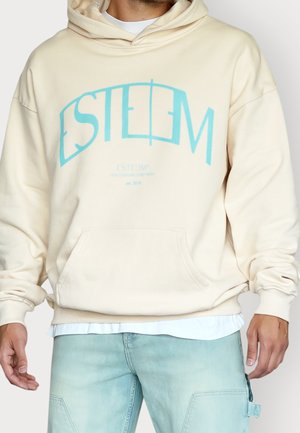 Mann trägt einen cremefarbenen Hoodie mit hellblauem "ESTEEM"-Schriftzug, weißem Unterhemd und hellblauen Jeans vor einfarbigem Hintergrund.