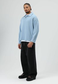 Hellblau übergroßes langärmeliges Polohemd mit Kragen, über einem weißen Hemd getragen, kombiniert mit schwarzen Jeans mit weitem Bein und schwarzen klobigen Schuhen.