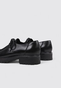 Zapatos oxford de cuero negro con punteras redondeadas, diseño de cordones y suelas de goma gruesas con patrones de tracción. Textura suave y detalles de costura.