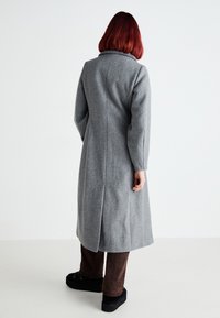 Gray wool coat with a high collar, long sleeves, a tailored fit, and a center back seam. Worn over brown pants and black platform shoes.