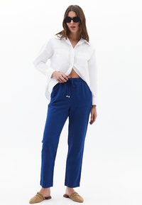 Witte button-up shirt met twee borstzakken, gecombineerd met blauwe broek met trekkoord. Model draagt zwarte zonnebril en beige instapschoenen.