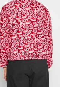 Rotes Langarmhemd mit weißem Paisley-Muster, getragen von einer Person, die steht, kombiniert mit schwarzen Hosen vor einem schlichten Hintergrund.