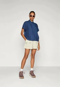 Woman wearing navy short-sleeve collared shirt, beige shorts with pockets, brown sneakers, white socks, and white sunglasses, standing with hands in pockets.