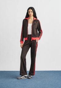 Καφέ αθλητική φόρμα με φωτεινές ροζ λεπτομέρειες, που περιλαμβάνει ζακέτα με φερμουάρ και ταιριαστό παντελόνι με τρεις κάθετες ρίγες. Υφασμένο από υφαντό ύφασμα.