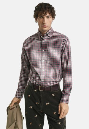 Camisa de cuadros en rojo y azul marino con un solo bolsillo en el pecho, combinada con pantalones de pana oscuros con bordados de aves.