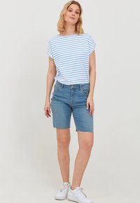Modra in bela črtasta kratka majica s kratkimi rokavi v kombinaciji s svetlo modrimi jeans kratkimi hlačami. Model nosi bele superge. Enobarvna ozadje.