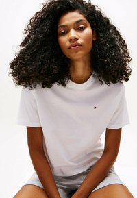 Witte katoenen t-shirt met een ronde halslijn en korte mouwen, voorzien van een klein logo op de borst. Model draagt grijze shorts.