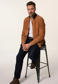 Bruine suède jas met een knoopsluiting aan de voorkant, zwarte kraag, gedragen over een wit shirt. Donkere jeans en bruin leren laarzen maken de look compleet.