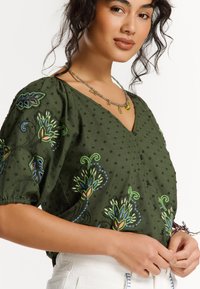 Blouse vert olive avec des manches bouffantes, ornée de motifs floraux brodés en bleu et vert, de boutons décoratifs et d'un décolleté en V.
