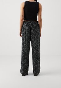 Kvinna klädd i svart ärmlös crop top och lösa svarta byxor med vitt geometriskt mönster, stående med ryggen mot en vit bakgrund.