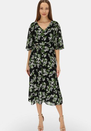 Kobieta ubrana w czarną sukienkę midi z białym kwiatowym i zielonym liściastym wzorem, dekolt w serek, rękawy do łokcia i beżowe sandały na wysokim obcasie, stojąca.