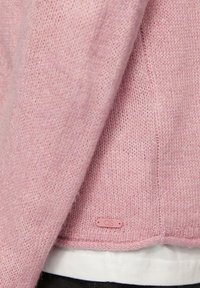 Lichtroze gebreide trui met een zachte textuur, voorzien van een klein QS-logo etiket nabij de zoom, gelaagd over een wit shirt.
