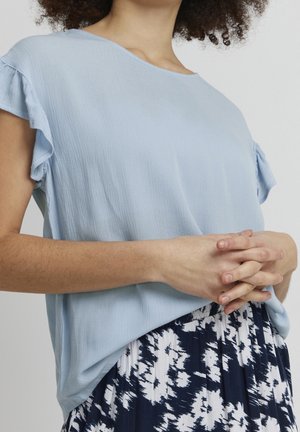 Kobieta ubrana w jasnoniebieską bluzkę z krótkim rękawem i falbankami oraz w czarno-białą wzorzystą spódnicę, ręce złożone na wysokości talii.