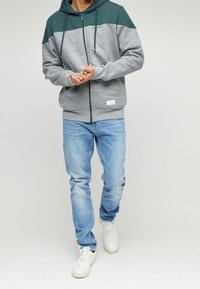 Mężczyzna w zielono-szarym rozpinanym hoodie, jasnoniebieskich dżinsach i białych tenisówkach stoi z rękami złożonymi. 