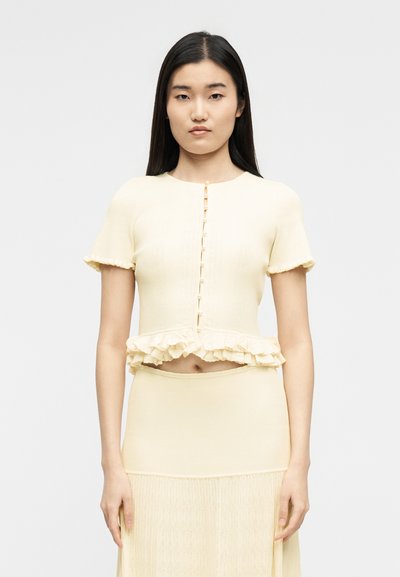 Γυναίκα με μακριά μαύρα μαλλιά, που φοράει απαλό κίτρινο κοντομάνικο πουκάμισο με κουμπιά και βολάν στο τελείωμα, και φούστα εποχιακή, στέκεται μπροστά σε έναν απλό φόντο.